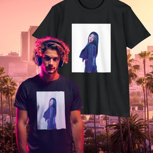 May include: Black t-shirts featuring a rectangular image of a person in a purple outfit. The person is standing and looking over their shoulder. The background is white. The t-shirts are displayed in front of a cityscape with palm trees.