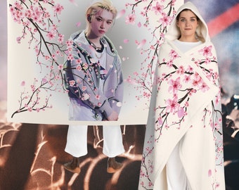 Puede incluir: Una manta blanca con capucha con estampado de flores de cerezo rosa. La manta presenta una foto de una persona con una chaqueta gris claro y cabello negro. La manta se muestra drapeada sobre una persona.