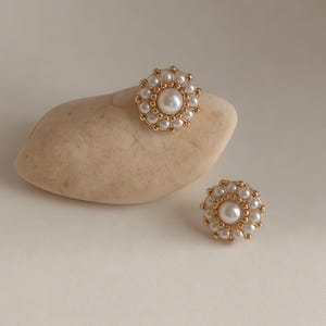 May include: Two gold-toned, pearl-studded earrings. Each earring features a large central pearl surrounded by smaller pearls, creating a floral design. One earring rests on a small, light-colored stone, while the other sits separately.