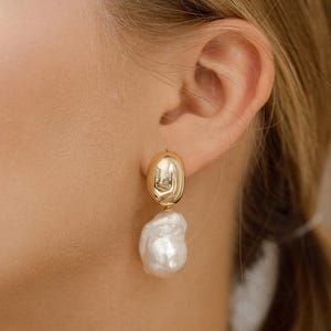 May include: Gold-tone earrings featuring a smooth, oval top and a large, irregular white pearl drop. The earrings are shown on a person's ear, highlighting their elegant design and contrasting textures.