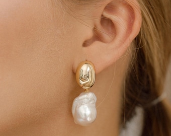 May include: Gold-tone earrings featuring a smooth, oval top and a large, irregular white pearl drop. The earrings are shown on a person's ear, highlighting their elegant design and contrasting textures.