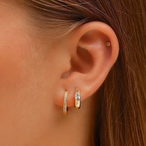 May include: A gold-toned ear stack consisting of two small hoop earrings and one tiny stud earring. One hoop features a row of small clear stones, while the other hoop has a single oval-shaped clear stone set in the center of the metal band.