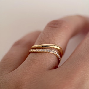 Puede incluir: Dos anillos dorados en un dedo. El anillo superior es una banda simple, mientras que el anillo inferior es una banda dorada con pequeñas piedras transparentes engastadas en la parte superior. Los anillos se usan juntos.
