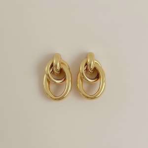 May include: A pair of gold-colored earrings. Each earring features a linked oval design with a smaller, rounded top element. The earrings have a polished, reflective surface, and are set against a neutral background.