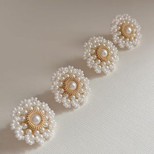 K&ouml;nnte beinhalten: Vier wei&szlig;e, perlenf&ouml;rmige Ohrringe in Blumenform mit goldfarbenen Zentren. Die Ohrringe sind diagonal auf einer wei&szlig;en Oberfl&auml;che angeordnet. Jeder Ohrring hat eine zentrale Perle, die von einem goldfarbenen Ring und wei&szlig;en Perlen umgeben ist.