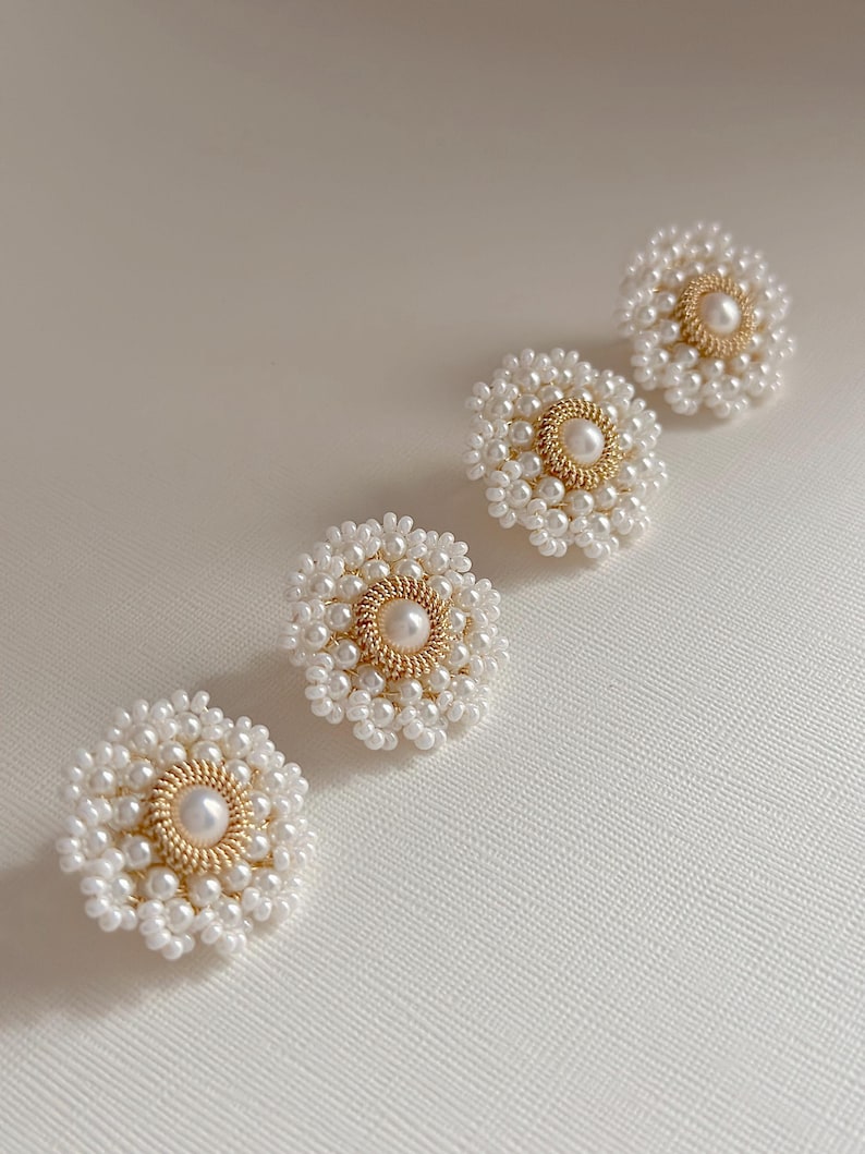 K&ouml;nnte beinhalten: Vier wei&szlig;e, perlenf&ouml;rmige Ohrringe in Blumenform mit goldfarbenen Zentren. Die Ohrringe sind diagonal auf einer wei&szlig;en Oberfl&auml;che angeordnet. Jeder Ohrring hat eine zentrale Perle, die von einem goldfarbenen Ring und wei&szlig;en Perlen umgeben ist.