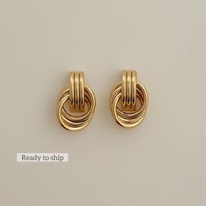 May include: A pair of gold-colored earrings. Each earring features a looped design with three vertical bars at the top and two interlocking oval shapes below. The earrings have a shiny finish and are displayed on a neutral background.