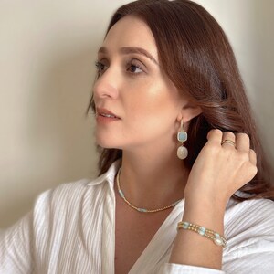 Pode incluir: Uma mulher vestindo uma camisa branca com um colar de corrente de ouro e uma pulseira de ouro com contas azuis e brancas. Ela tamb&eacute;m est&aacute; usando um par de brincos de ouro com pedras azuis e brancas.