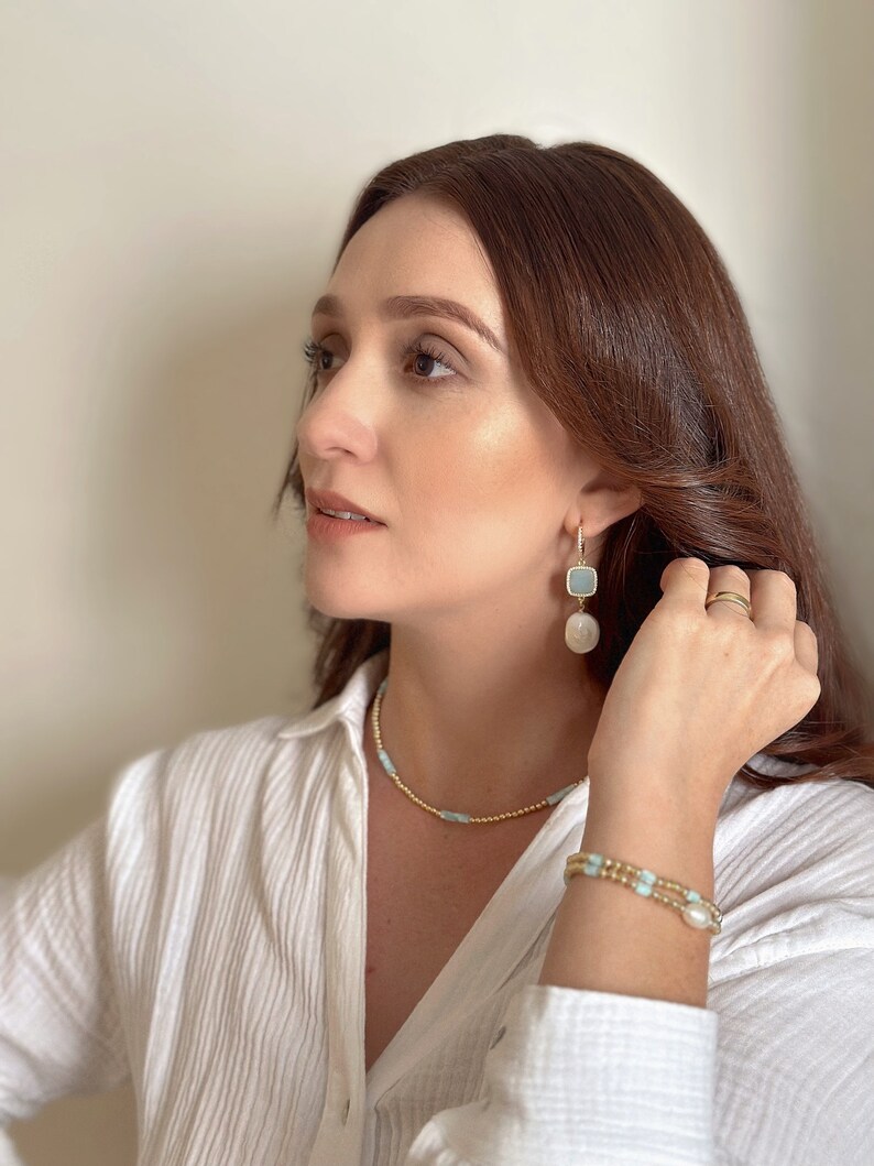 Pode incluir: Uma mulher vestindo uma camisa branca com um colar de corrente de ouro e uma pulseira de ouro com contas azuis e brancas. Ela tamb&eacute;m est&aacute; usando um par de brincos de ouro com pedras azuis e brancas.