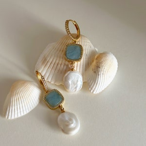 Pode incluir: Um par de brincos de ouro com uma pedra preciosa azul e uma p&eacute;rola branca pendente. Os brincos s&atilde;o exibidos em um fundo branco com conchas.