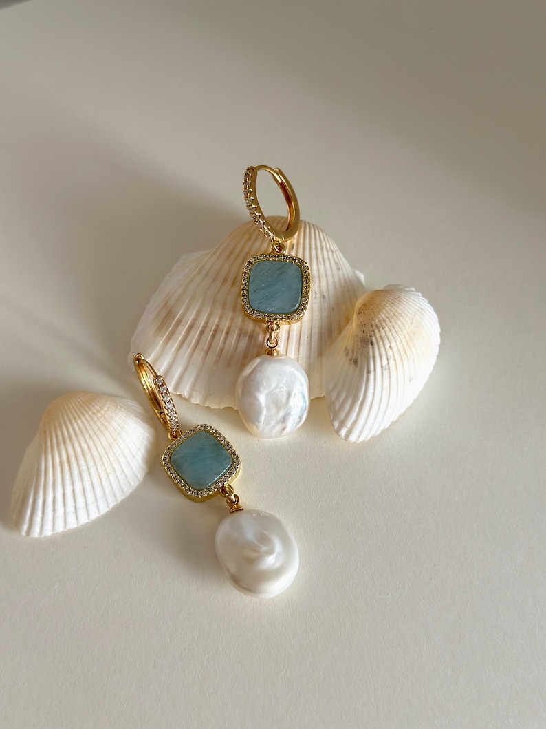 Pode incluir: Um par de brincos de ouro com uma pedra preciosa azul e uma p&eacute;rola branca pendente. Os brincos s&atilde;o exibidos em um fundo branco com conchas.