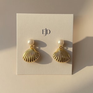 May include: Gold-colored seashell earrings with a pearl accent. The earrings are displayed on a white card with the initials "EJD" printed in black. The earrings have a textured, ridged design.