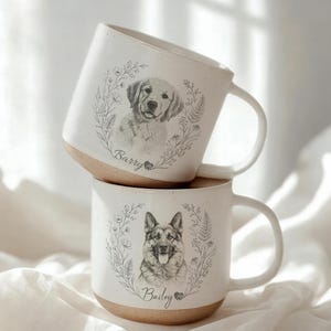 Peut inclure: Deux mugs en céramique blanche empilés avec une base marron clair. Chaque mug présente un croquis détaillé d'un chien entouré d'une couronne florale. Le mug du haut a un Golden Retriever, celui du bas un Berger Allemand. Noms "Barry" et "Bailey".