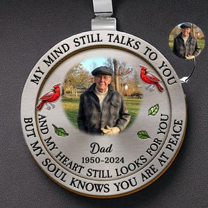Puede incluir: Un adorno circular plateado para coche con una foto de un hombre con sombrero. El adorno tiene el texto "MY MIND STILL TALKS TO YOU" y "MY HEART STILL LOOKS FOR YOU". También tiene el texto "Dad 1950-2024". También aparecen dos cardenales rojos y hojas verdes.