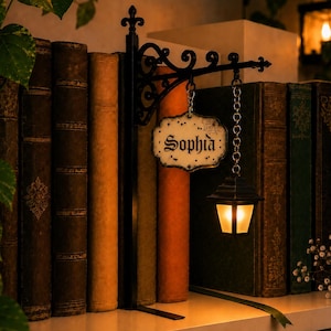 May include: A black metal bookend featuring an ornate bracket that holds a hanging sign with the name Sophia in black gothic lettering and a small, illuminated lantern suspended by a silver chain, positioned between several vintage-style books.