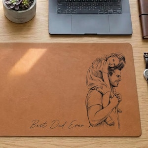 May include: A rectangular, tan leather desk mat with a black sketch of a father carrying a child on his shoulders. The text "Best Dad Ever" is printed in black. A laptop, pen, and succulent plant are also visible.