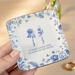 Op de afbeelding: Vierkante, wit en blauw bloemen sieraden schaal met gouden rand. De schaal heeft een blauw bloemenontwerp met de namen "Sylvia" en "Sophie" en de quote "It takes a long time to grow an old friend."