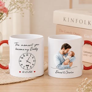Op de afbeelding: Twee witte keramische mokken met rode handvatten. Eén mok heeft een klokontwerp met de tekst "The moment you become my Daddy" en de datum 02-23-2026. De andere mok heeft een illustratie van een vader die een baby vasthoudt, met de tekst "David & Charlie".