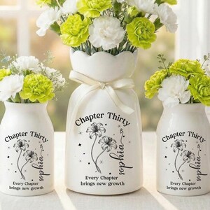 May include: Three white ceramic vases with floral arrangements. The vases are decorated with the text "Chapter Thirty", floral illustrations, and the name "Sophia". The flowers are white and lime green carnations. A white ribbon adorns the largest vase.