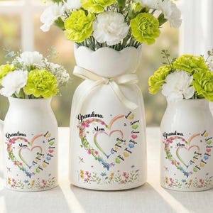 May include: Three white ceramic vases with floral arrangements. The vases are decorated with the words "Grandma" and "Mom" inside a heart-shaped design. The flowers are white and green.