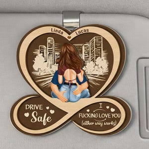 Op de afbeelding: Houten auto-accessoire met een hartvormig ontwerp. Toont een knuffelend stel, een stadsgezicht en de zinnen "Drive Safe" en "I Fucking Love You (either way works)". De namen "Linda" en "Lucas" zijn ook aanwezig.
