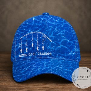 Op de afbeelding: Blauwe baseballcap met een waterachtig patroon. De cap heeft een wit hengelontwerp met de woorden "REEL COOL GRANDPA" en namen. Een klein logo is rechtsonder zichtbaar.