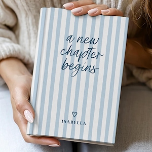 Puede incluir: Un diario de tapa dura con un patrón de rayas azul claro y blanco. La portada presenta el texto "a new chapter begins" en una fuente manuscrita, un pequeño corazón y el nombre "ISABELLA". El diario está en manos de alguien.