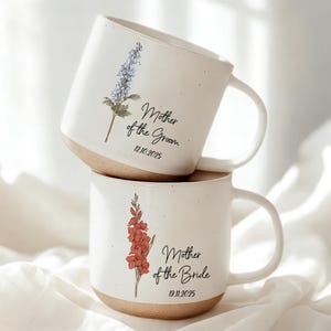 May include: Two white ceramic mugs stacked on top of each other. Each mug features a floral design and the text "Mother of the Groom" and "Mother of the Bride" with dates. The mugs have a speckled finish and a tan base.