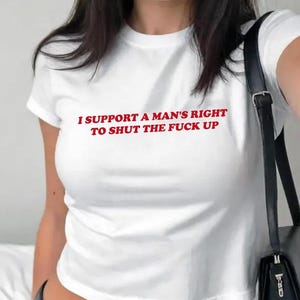 Può includere: Top corto bianco con la scritta rossa: "I SUPPORT A MAN'S RIGHT TO SHUT THE FUCK UP." La maglietta ha maniche corte e scollo tondo. Una borsa nera è visibile sul lato destro dell'immagine.