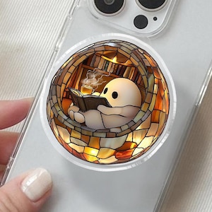 May include: A clear phone case with a pop-up grip featuring a cartoon figure reading a book inside a stained-glass window design. The pop-up grip has a circular shape with warm colors, including yellow, orange, and brown.