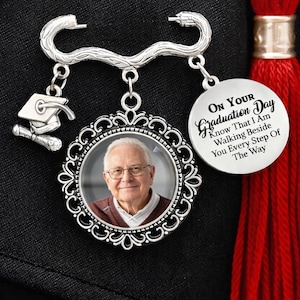 Könnte beinhalten: Ein silberfarbener Abschluss-Pin mit einem Foto eines Mannes in einem dekorativen Rahmen, einem Mortarboard-Anhänger und einer Scheibe mit dem Text "ON YOUR Graduation Day Know That I Am Walking Beside You Every Step Of The Way". Eine rote Quaste ist sichtbar.