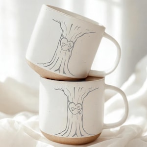 Peut inclure: Deux mugs en céramique blanche empilés. Chaque mug présente un motif d'arbre gris avec un emblème en forme de cœur portant les initiales "D+S". Le bas de chaque mug est de couleur beige naturel, et chacun a une anse blanche.