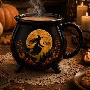 May include: A black ceramic cauldron-shaped mug with three small feet and a handle. The front features a graphic of a witch flying on a broomstick in front of a large yellow moon, surrounded by autumn leaves and the name Olivia printed in black.