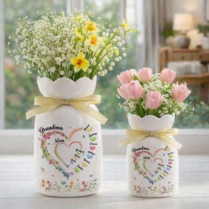 May include: Two white ceramic vases with floral arrangements. The vases have a scalloped top and a gold ribbon. The larger vase has the words "Grandma Mom" with a heart design and names. The smaller vase has the same design. One vase has yellow flowers, the other has pink tulips.