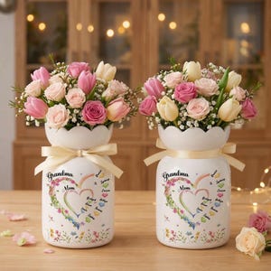 May include: Two white ceramic vases with floral arrangements. Each vase has a cream-colored ribbon and features the words "Grandma" and "Mom" with a heart-shaped floral design. The arrangements include pink roses, tulips, and white flowers.