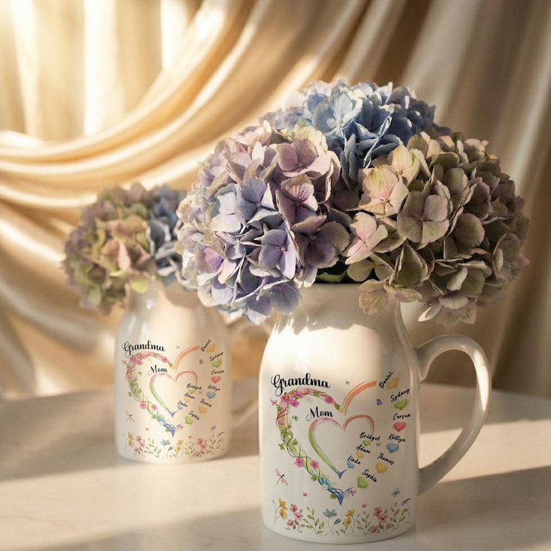 May include: Two white ceramic vases with hydrangea flowers. The vases are decorated with a heart-shaped design that says "Grandma Mom" and includes names. The flowers are a mix of blue, purple, and green.