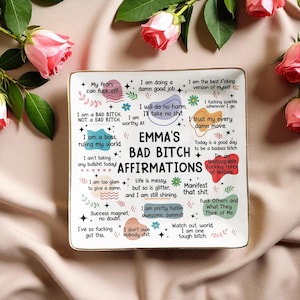 May include: A rectangular ceramic tray with a gold rim, featuring the text "EMMA'S BAD BITCH AFFIRMATIONS" in black. The tray is decorated with various affirmations in different fonts and colors, including phrases like "I am a boss" and "Manifest that shit."