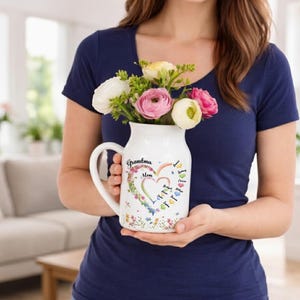May include: A white ceramic pitcher with a handle, filled with colorful flowers. The pitcher is decorated with a heart-shaped design, the words "Grandma Mom", and names. The flowers include white, pink, and yellow blooms. The pitcher is held by a person wearing a navy blue shirt.