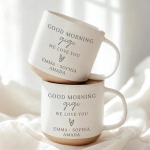 May include: Two white ceramic mugs stacked on top of each other. Each mug has a light brown base and a curved handle. The text "GOOD MORNING gigi WE LOVE YOU" is printed on the front, along with the names "EMMA | SOPHIA AMADA".
