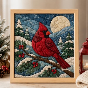 Op de afbeelding: Een vierkant ingelijst wandkunstwerk van stof met een doorgestikte scène van een felrode kardinaal op een dennentak met rode bessen. De achtergrond toont een besneeuwd winterlandschap met groenblijvende bomen en een grote bleke maan in de lucht.