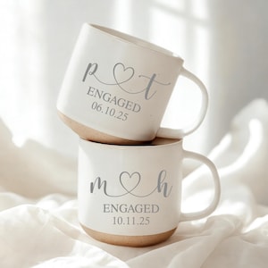 Può includere: Due tazze in ceramica bianca impilate. Ogni tazza ha una base marrone chiaro e un manico curvo. La tazza superiore ha le lettere "p♡t" e il testo "ENGAGED 06.10.25" stampati in grigio. La tazza inferiore ha le lettere "m♡h" e il testo "ENGAGED 10.11.25" stampati in grigio.