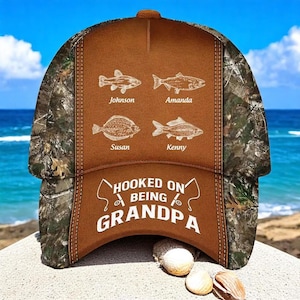 May include: A brown and camouflage baseball cap with the text "Hooked on Being Grandpa." The cap features four fish illustrations with names above them. The cap is sitting on a sandy surface.