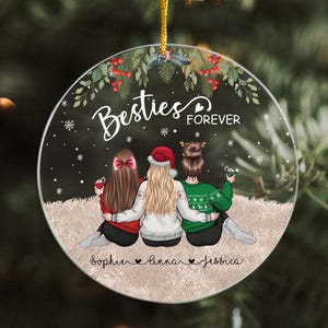 Pu&ograve; includere: Un ornamento natalizio rotondo con tre figure, ognuna con un'acconciatura diversa e che indossa un maglione rosso, un cappello da Babbo Natale e un maglione verde. L'ornamento recita "Besties Forever" con i nomi sotto.