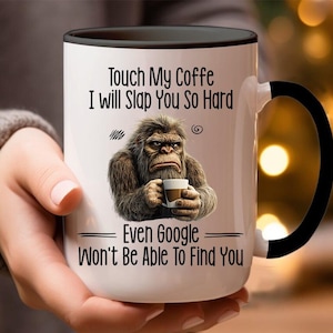 Op de afbeelding: Een witte keramische koffiemok met een zwarte handgreep. De mok heeft een cartoonillustratie van een Bigfoot die een kopje koffie vasthoudt. De tekst op de mok luidt "Touch My Coffee I Will Slap You So Hard Even Google Won't Be Able To Find You."