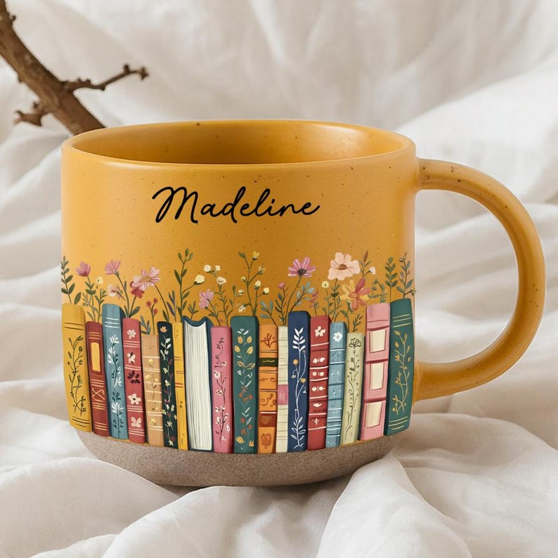May include: Mustard yellow ceramic mug with a wraparound design of colorful books and flowers. The mug has a tan base and a large handle. The name "Madeline" is written in black script on the front of the mug.