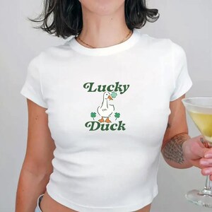 May include: White cropped t-shirt with a green and white cartoon duck holding a shamrock and the text "Lucky Duck" in green.