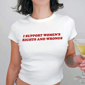 May include: White crop top with the red text "I SUPPORT WOMEN'S RIGHTS AND WRONGS". The shirt is a short-sleeved, fitted style. A martini glass with a green olive is in the background.