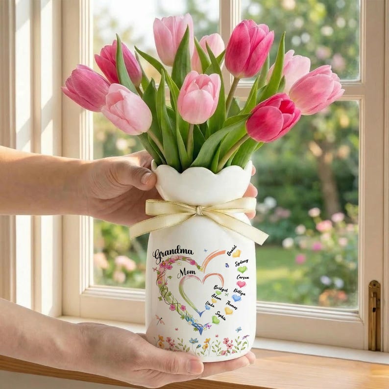 May include: A white ceramic vase with a scalloped rim, tied with a beige ribbon, holds a bouquet of pink tulips. The vase is decorated with the words "Grandma Mom" and a heart-shaped floral design, with names listed on the side.