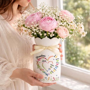 May include: A white ceramic vase with a floral arrangement of pink peonies and white baby's breath. The vase is decorated with a heart-shaped design, the words "Grandma Mom", and names. A cream-colored ribbon is tied around the vase.