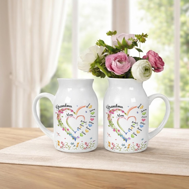 May include: Two white ceramic vases with handles, each adorned with a floral heart design and the words "Grandma" and "Mom." The vases are filled with colorful flowers and sit on a neutral-toned surface.